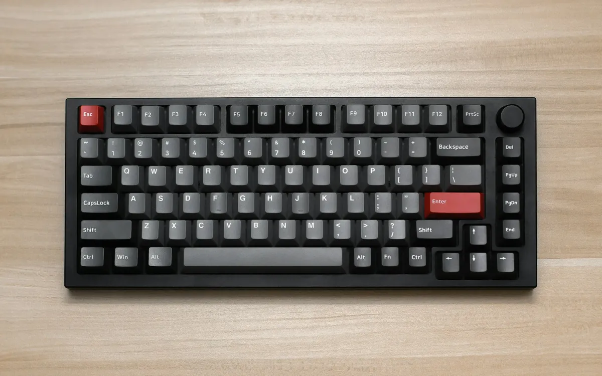A black 75% mechanical keyboard with red accent keys on a light wooden desk