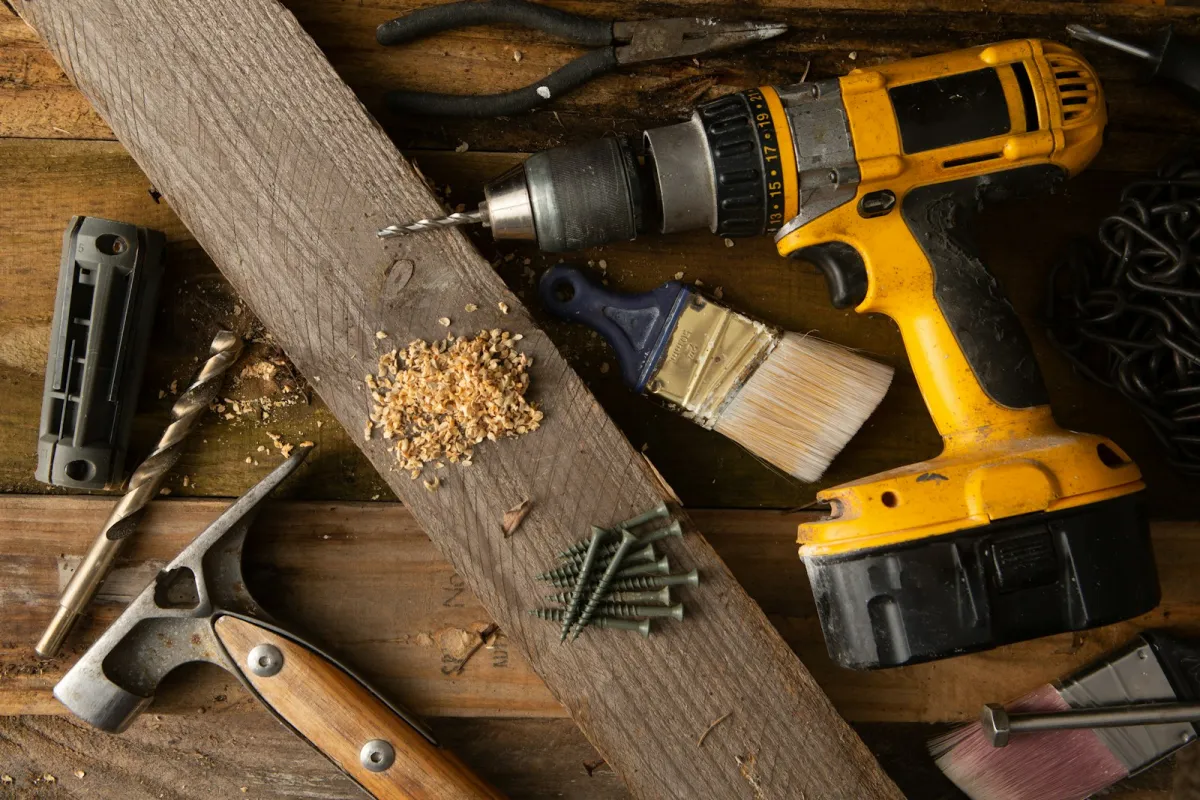 A cordless drill, hammer, pliers, paintbrush, drill bit and screws on a rough wooden workbench