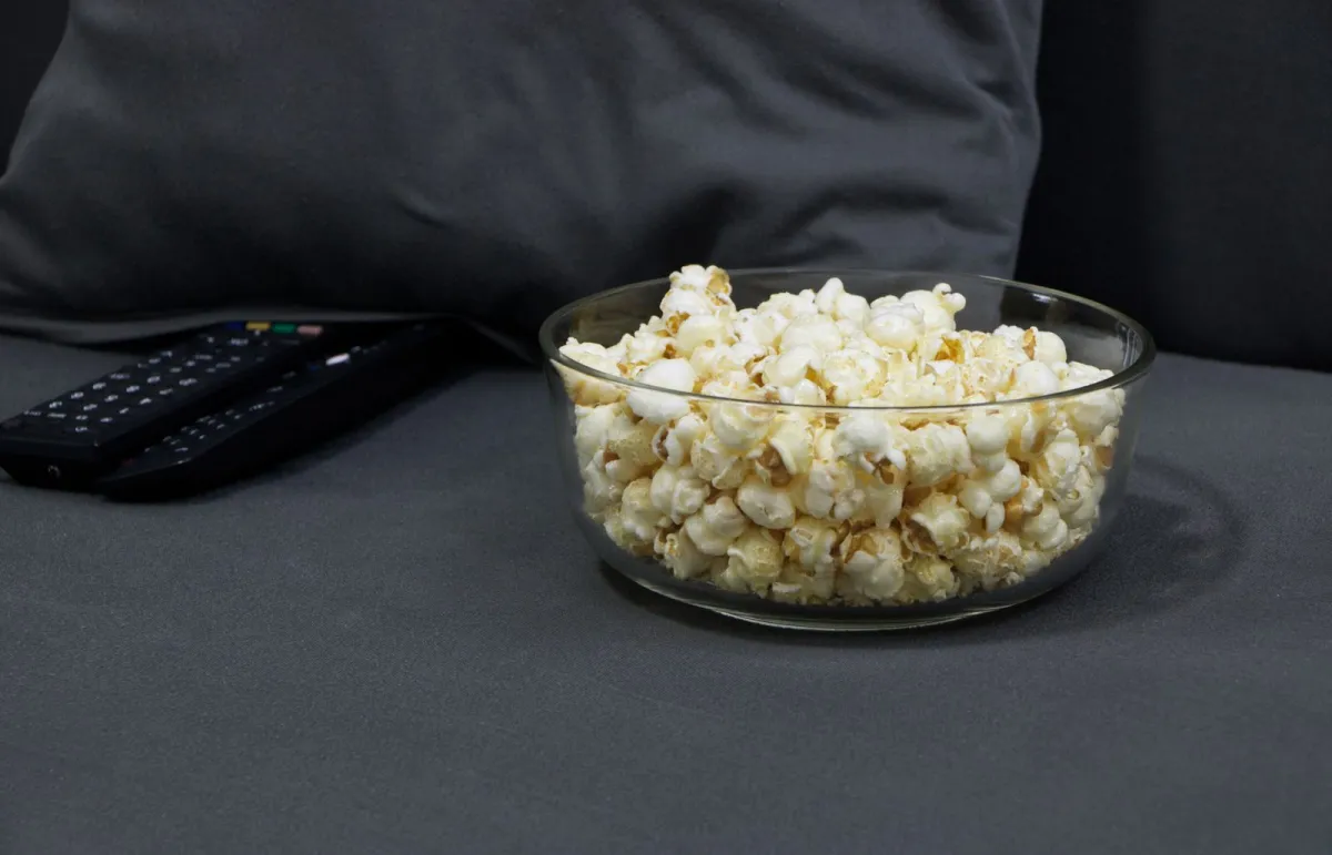 A glass bowl of popcorn next to two TV remote controls on a grey sofa A glass bowl of popcorn next to two TV remote controls on a grey sofa