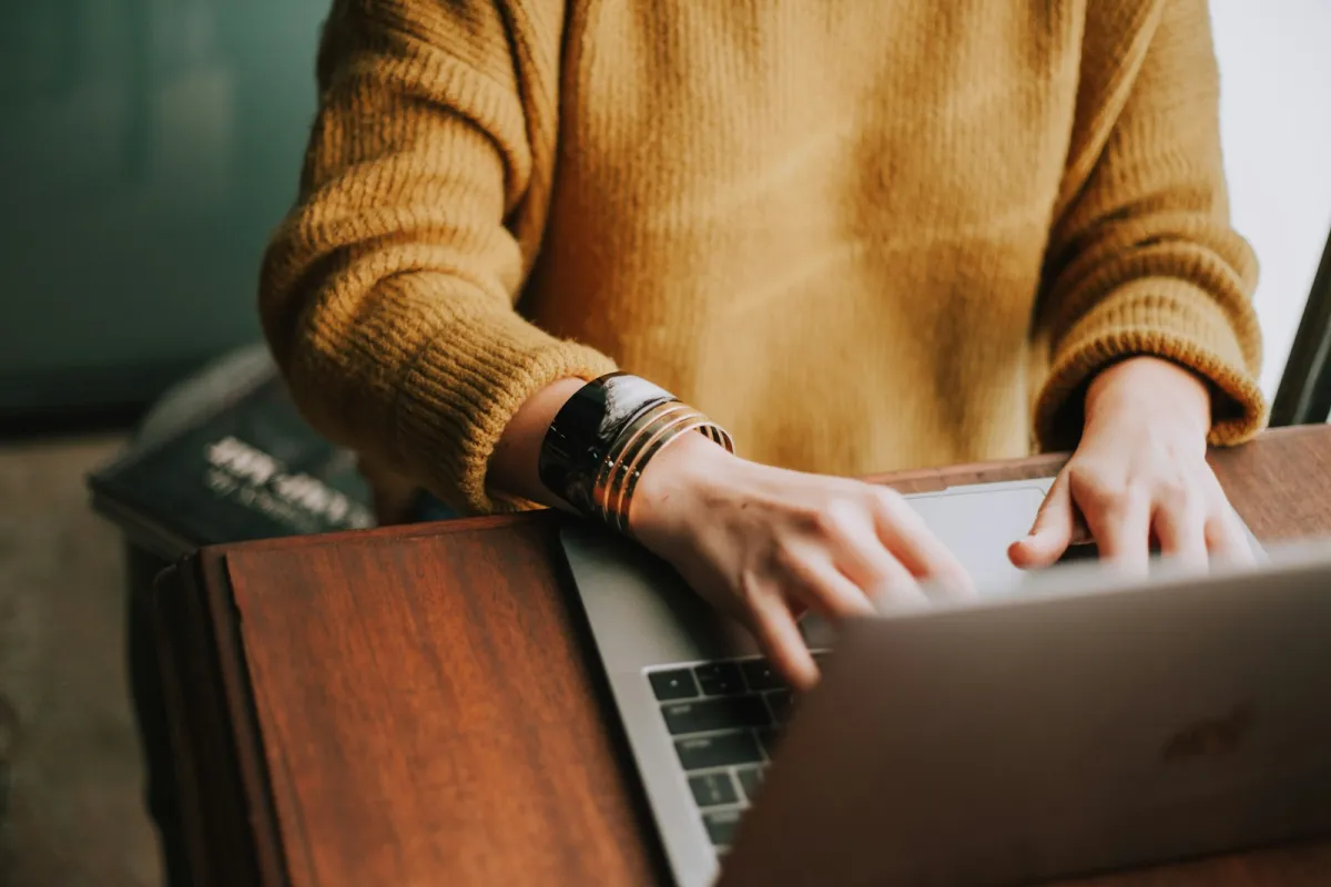 Person in a mustard jumper typing on a laptop at a wooden desk Person in a mustard jumper typing on a laptop at a wooden desk