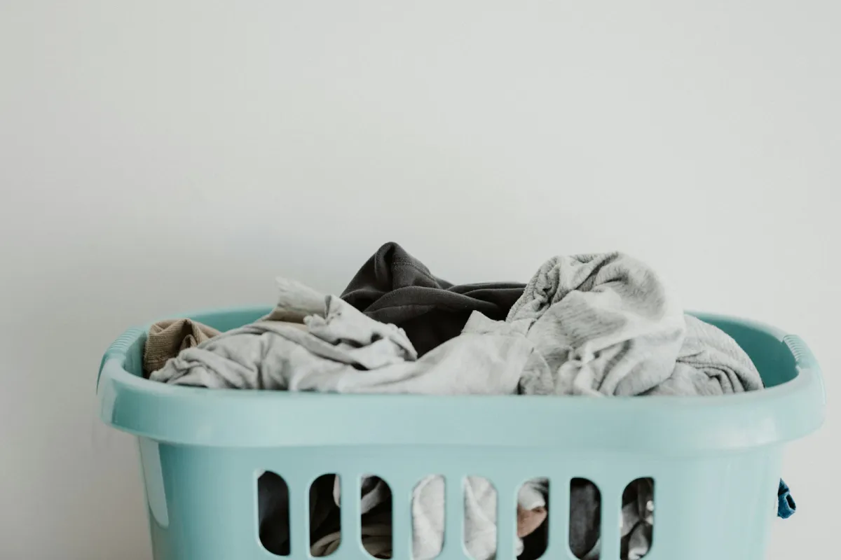 A pale blue plastic laundry basket full of grey and dark clothes against a white wall A pale blue plastic laundry basket full of grey and dark clothes against a white wall