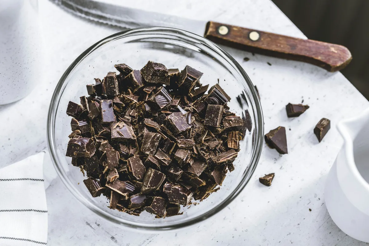 A glass bowl of chopped dark chocolate on a white table beside a knife