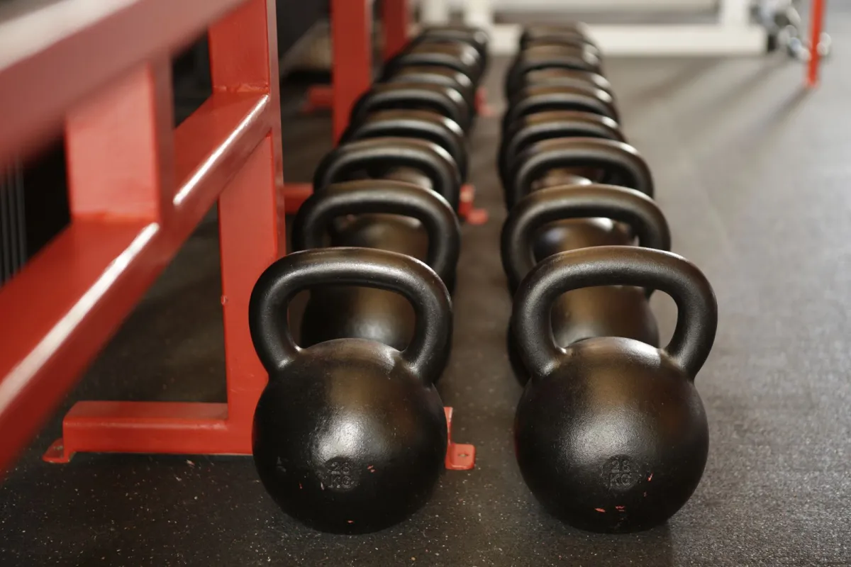 Two rows of black kettlebells lined up beside a red rack on a gym floor Two rows of black kettlebells lined up beside a red rack on a gym floor