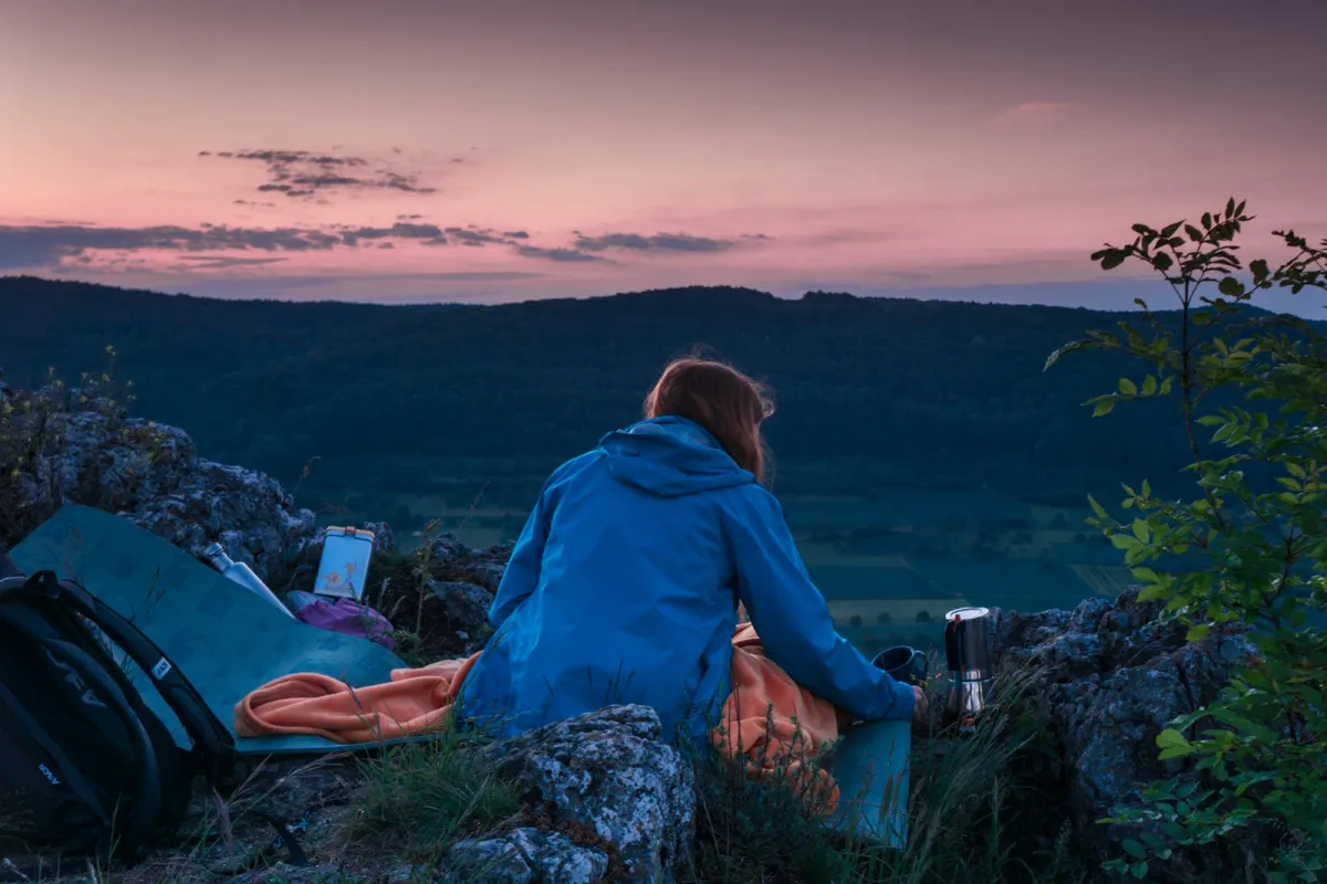 A camper with a flask of coffee looking out over hills at dusk