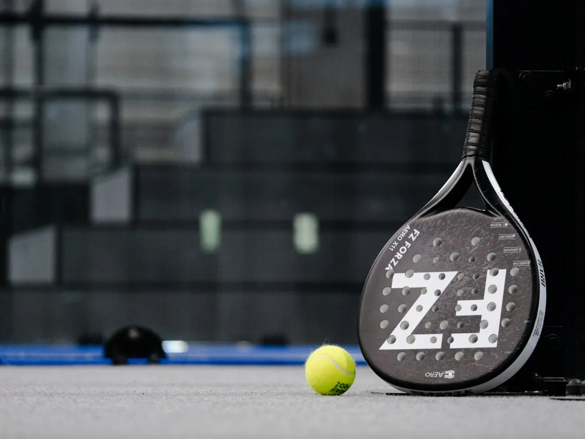 A padel racket and a yellow ball on a grey court surface A padel racket and a yellow ball on a grey court surface