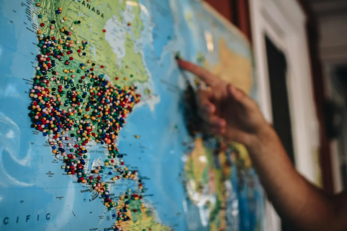 A hand pointing at a world map covered in coloured pins