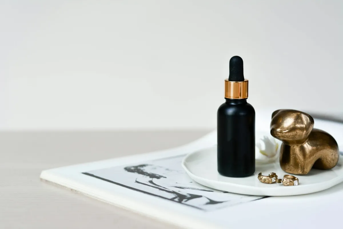 A black dropper bottle of serum on a white dish beside gold earrings