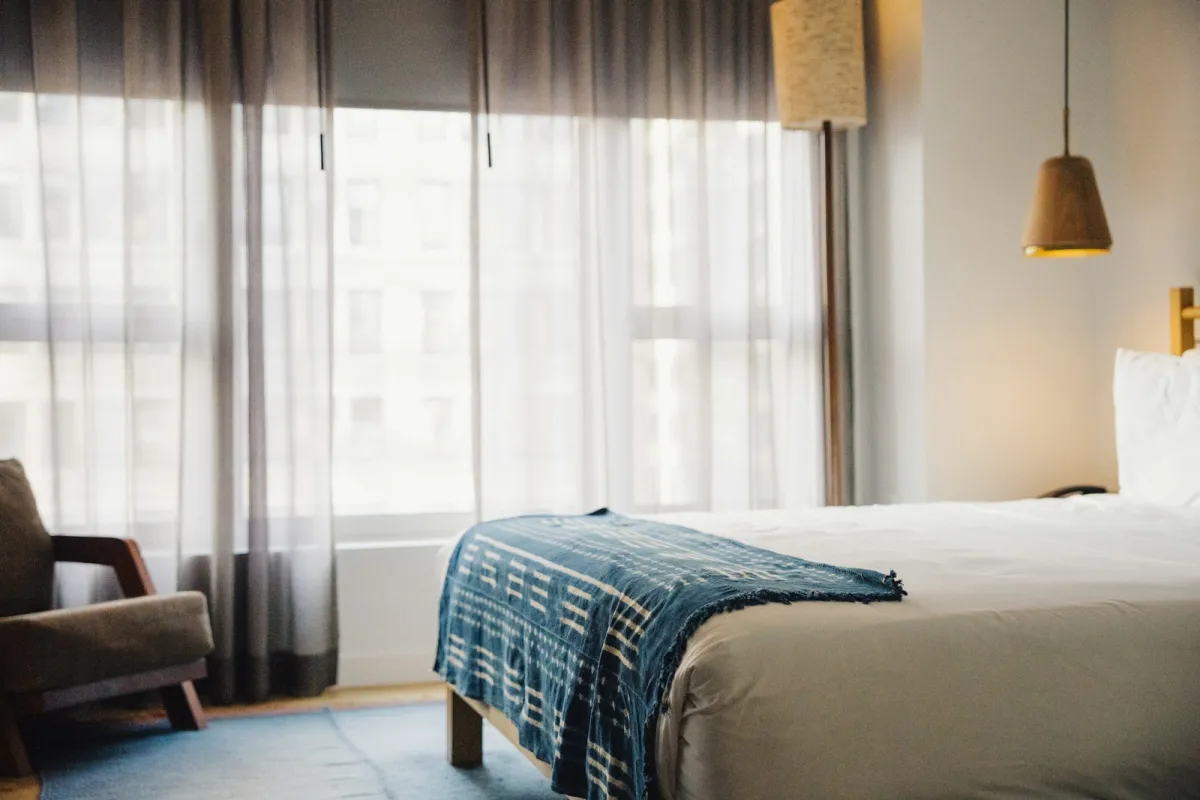 Bright hotel room with a made-up bed, a blue throw and sheer curtains Bright hotel room with a made-up bed, a blue throw and sheer curtains