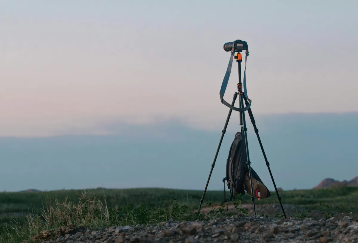A camera with a telephoto lens on a tripod in open grassland at dusk, a backpack hanging below