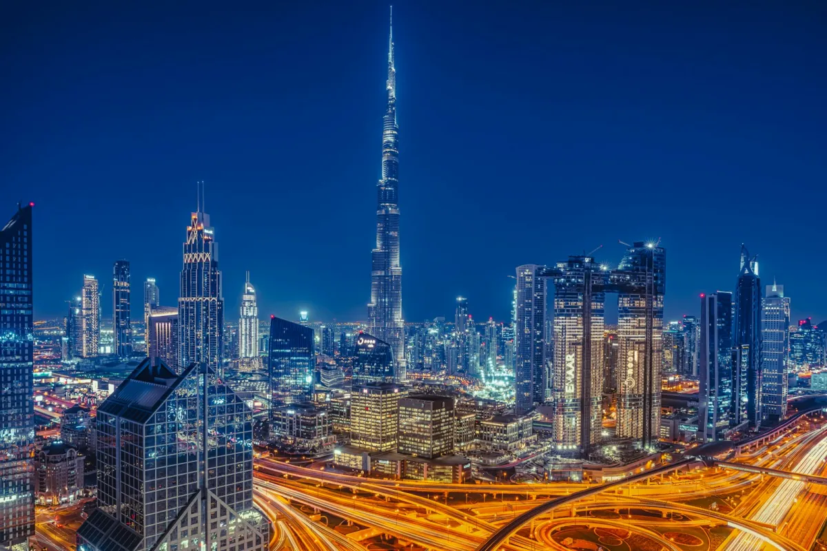 The Dubai skyline lit up at night with a tall tower in the centre and busy highways below