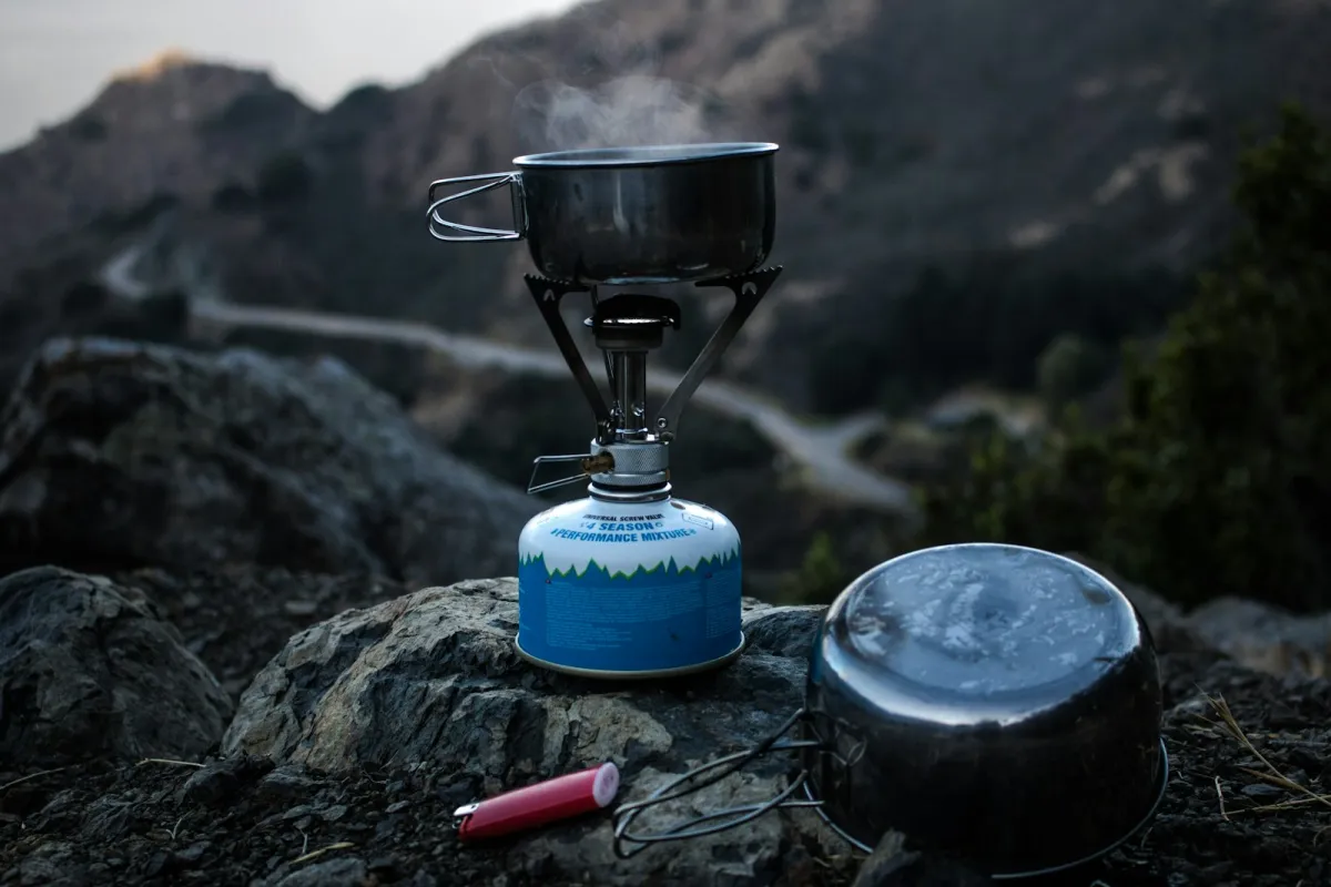 A camping stove heating a pot of water on a rocky hillside at dusk