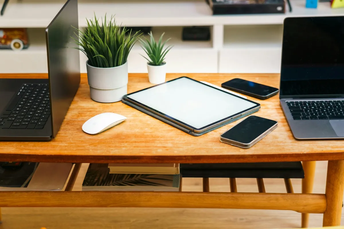 Two laptops, a tablet and two smartphones on a wooden desk with potted plants