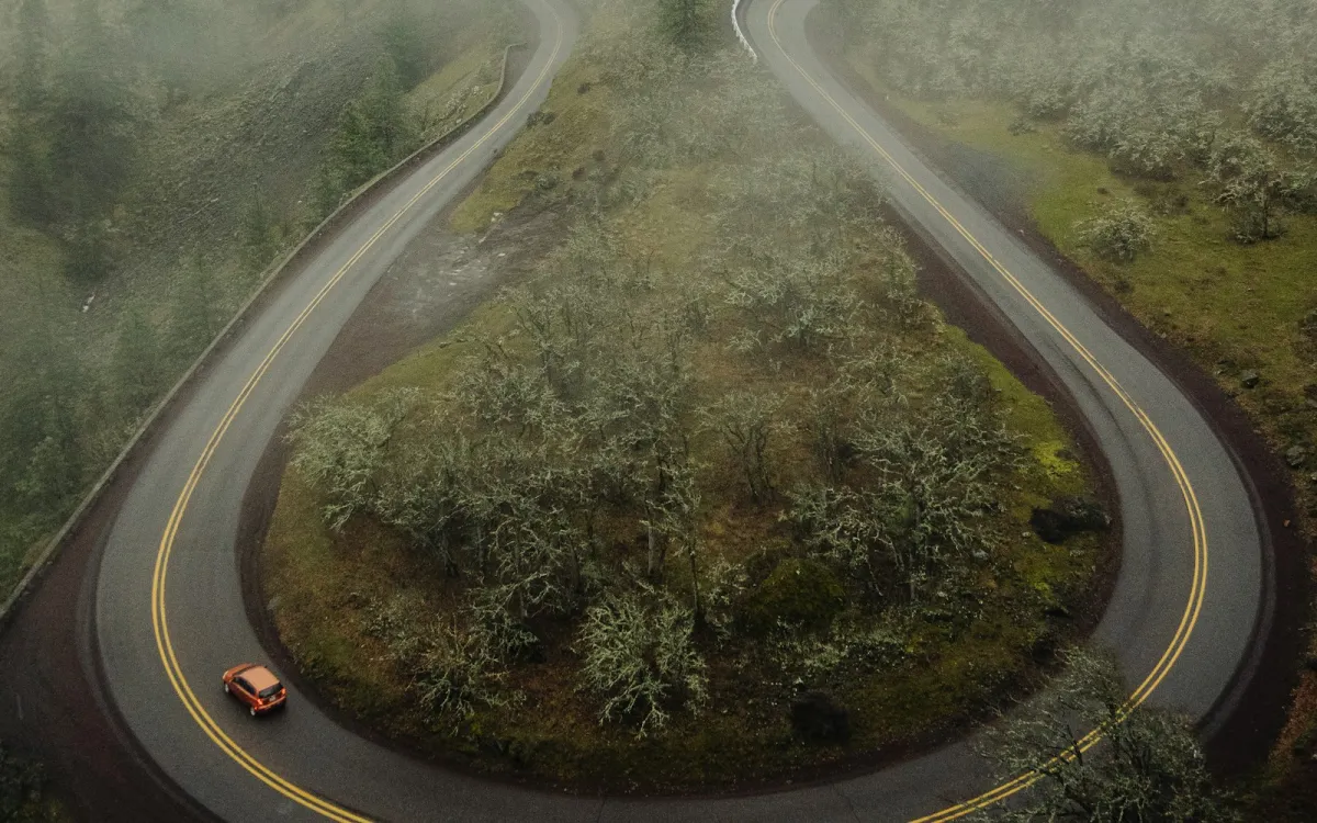 An orange car rounding a hairpin bend on a misty, tree-lined mountain road An orange car rounding a hairpin bend on a misty, tree-lined mountain road