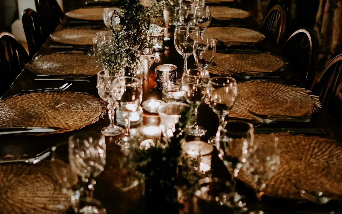 A long candlelit dining table laid with clear wine glasses, woven placemats and small flowers