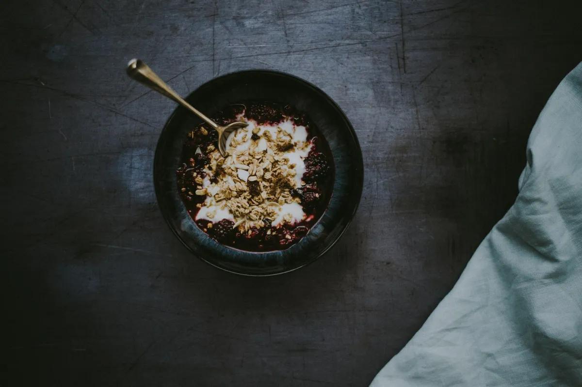 A bowl of yoghurt topped with granola and blackberries on a dark wooden table A bowl of yoghurt topped with granola and blackberries on a dark wooden table