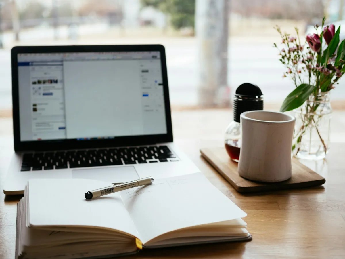 An open notebook and pen in front of a laptop on a wooden desk