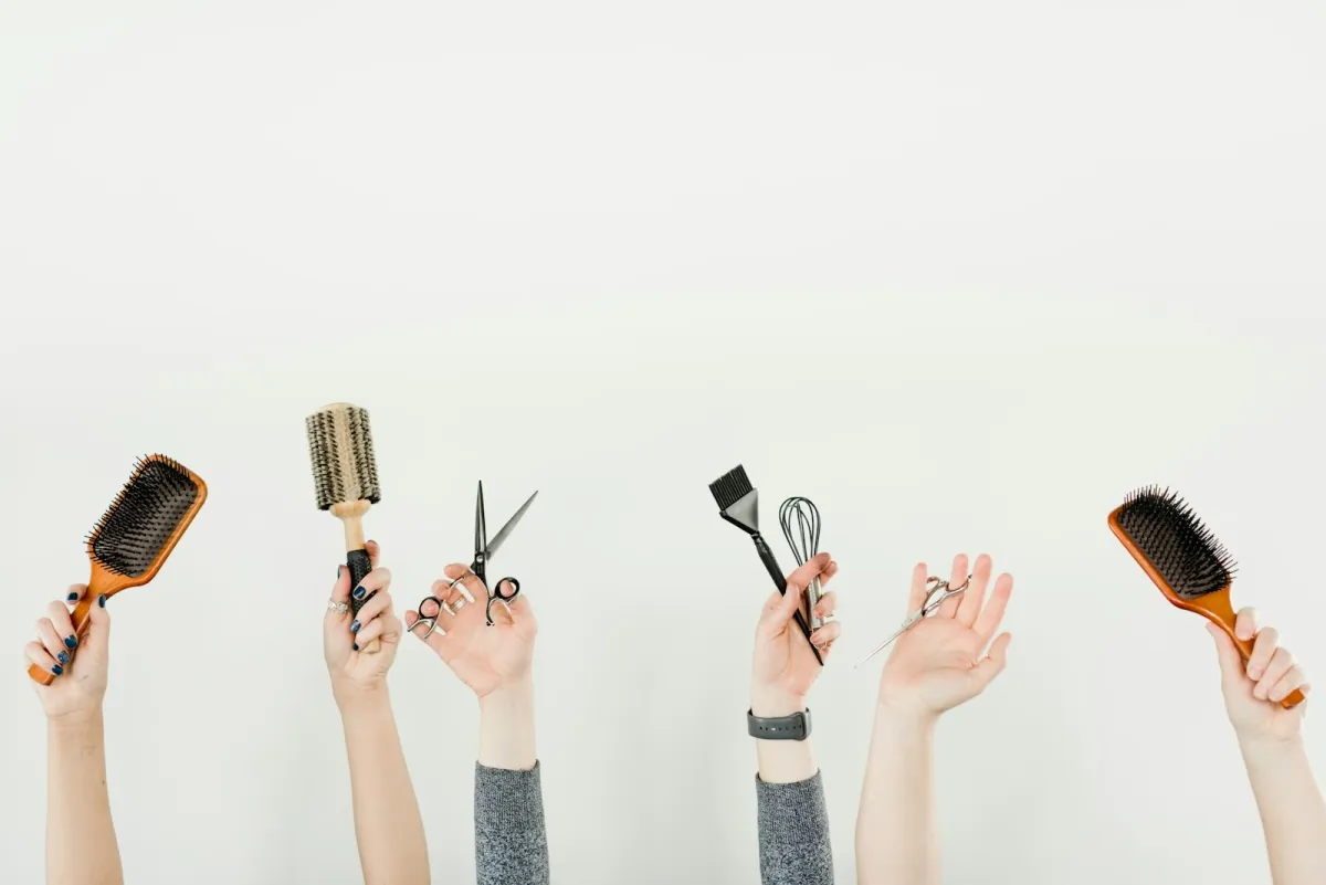 Raised hands holding hairbrushes, scissors and a tint brush against a white wall Raised hands holding hairbrushes, scissors and a tint brush against a white wall