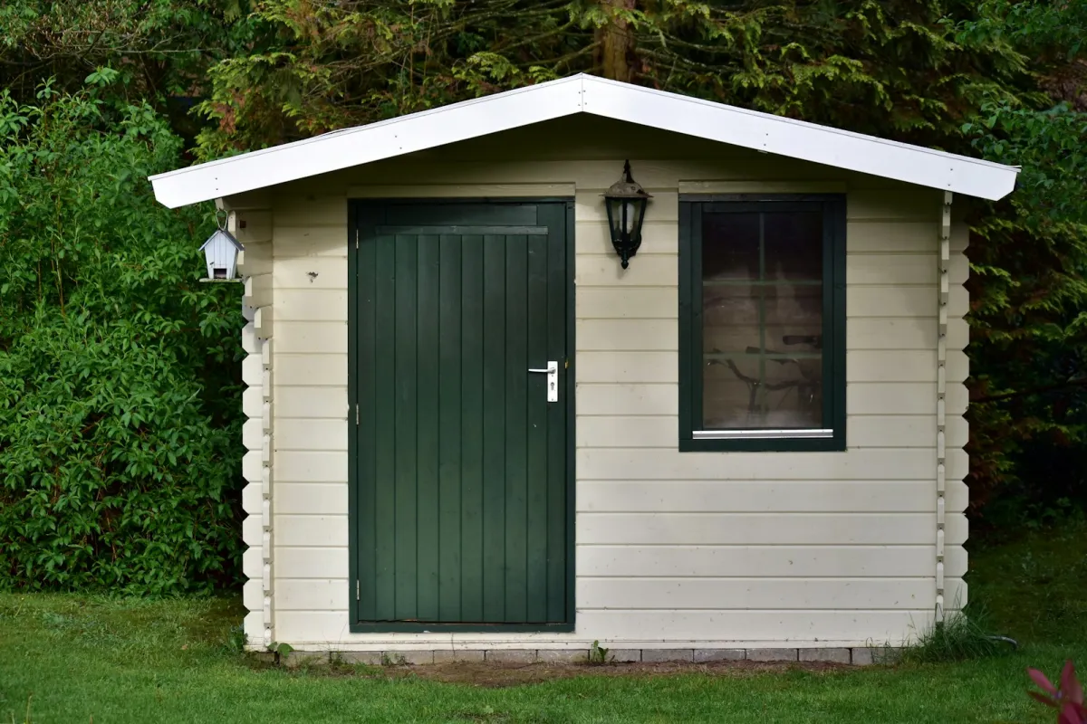 A small cream log cabin with a green door and window in a leafy garden