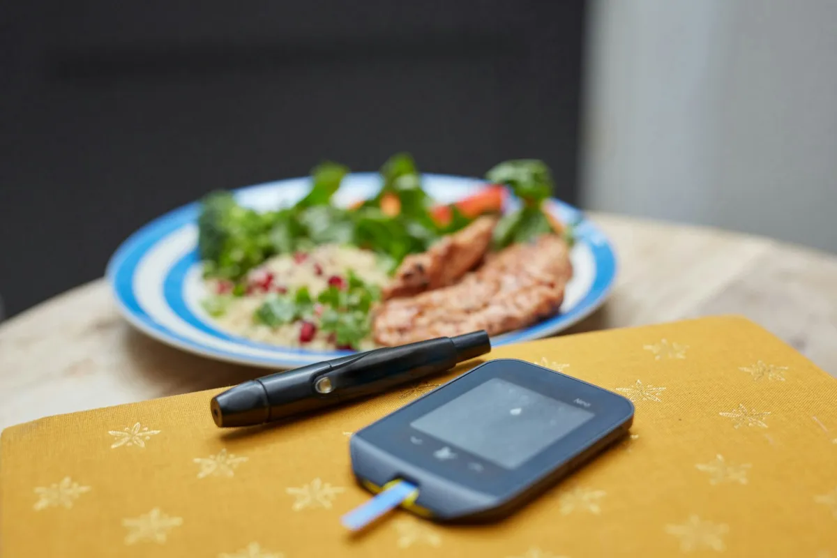 Blood glucose meter and lancing pen on a table in front of a plate of salad and chicken Blood glucose meter and lancing pen on a table in front of a plate of salad and chicken
