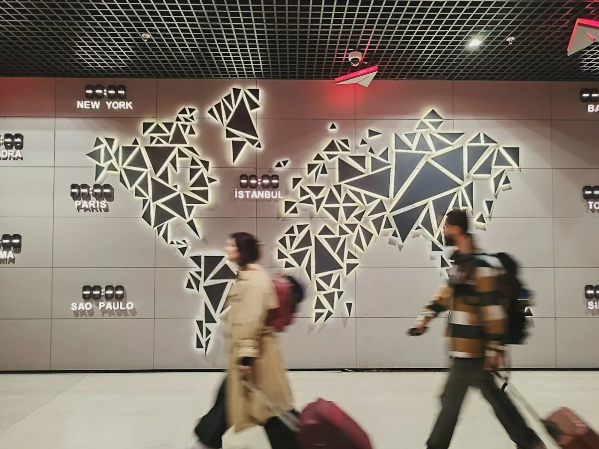 Two travellers with suitcases walk past a lit world map with time-zone clocks in an airport Two travellers with suitcases walk past a lit world map with time-zone clocks in an airport