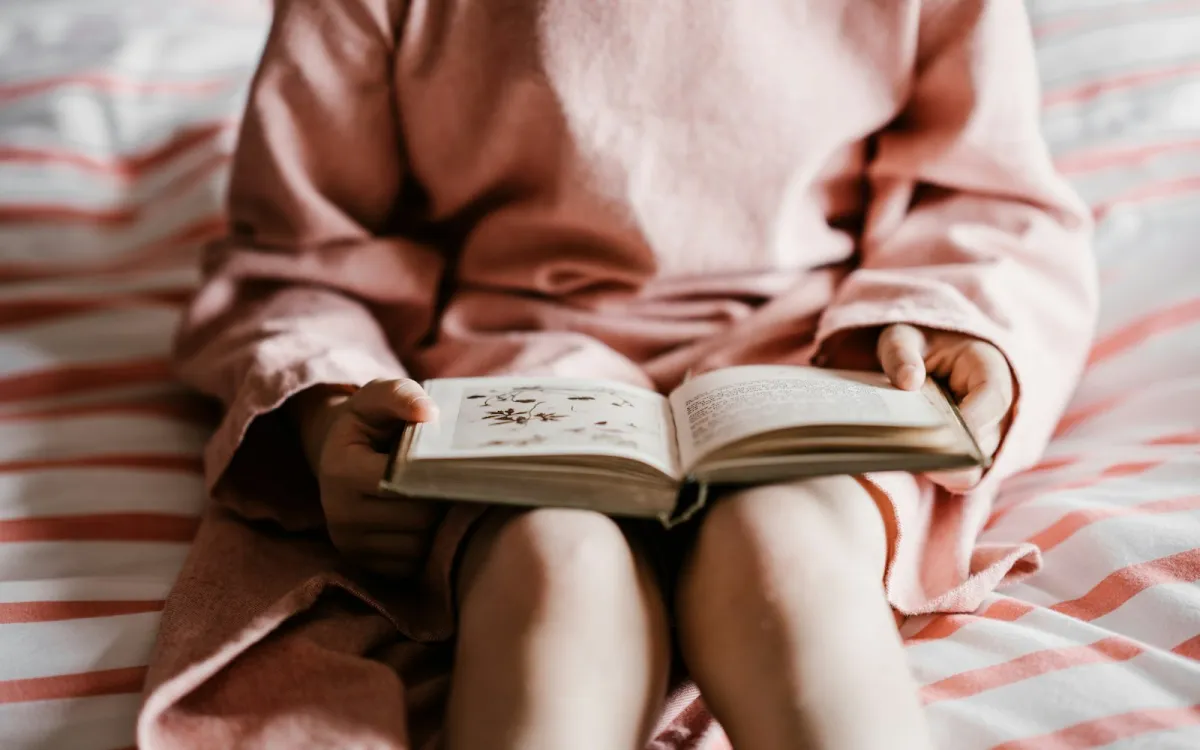A child in a pink dress reading an open book on a red and white striped bedspread A child in a pink dress reading an open book on a red and white striped bedspread