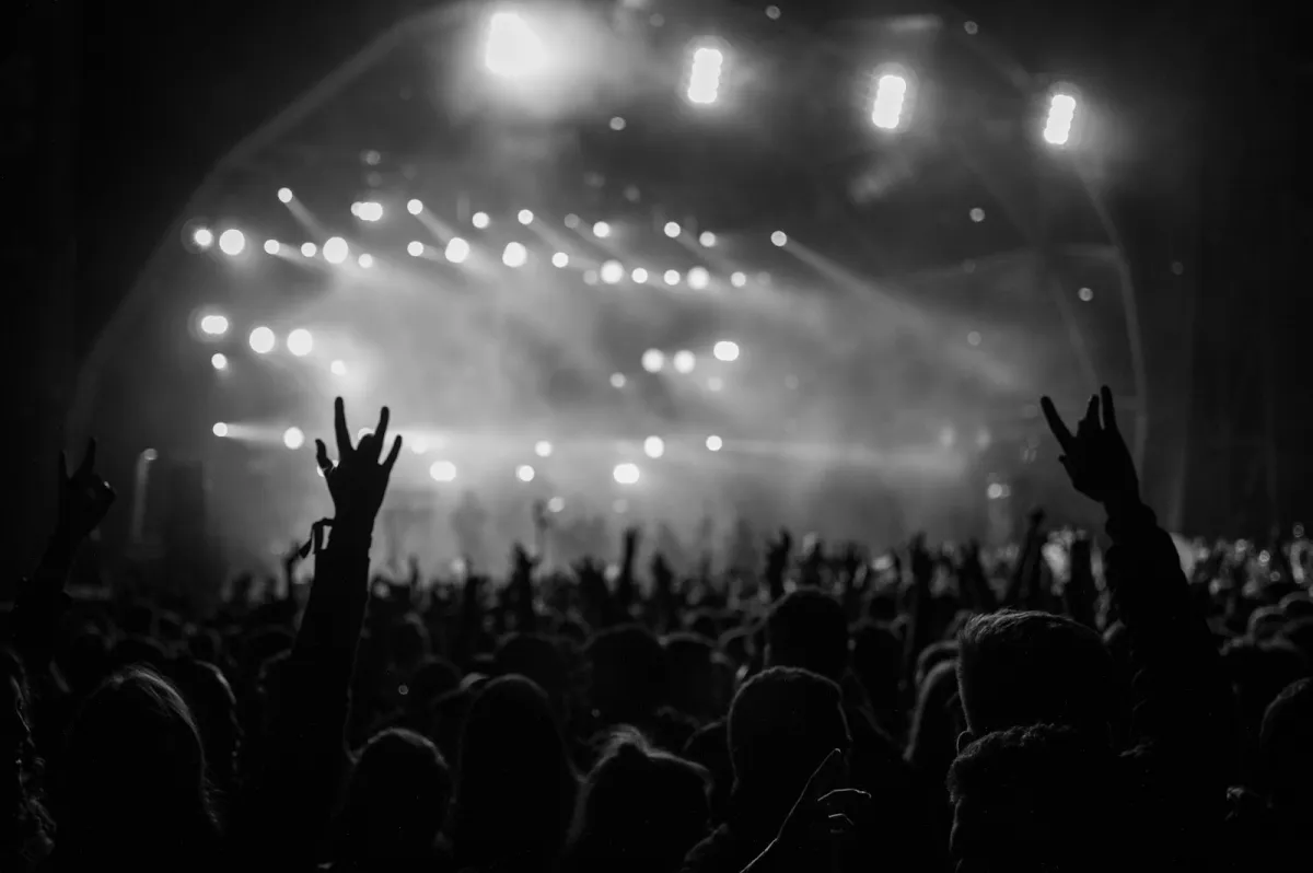 A black and white shot of a festival crowd with raised hands in front of a brightly lit stage A black and white shot of a festival crowd with raised hands in front of a brightly lit stage