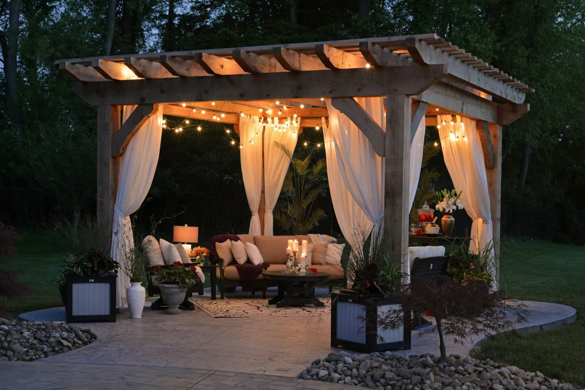 Wooden pergola with curtains and string lights over a garden seating area at dusk