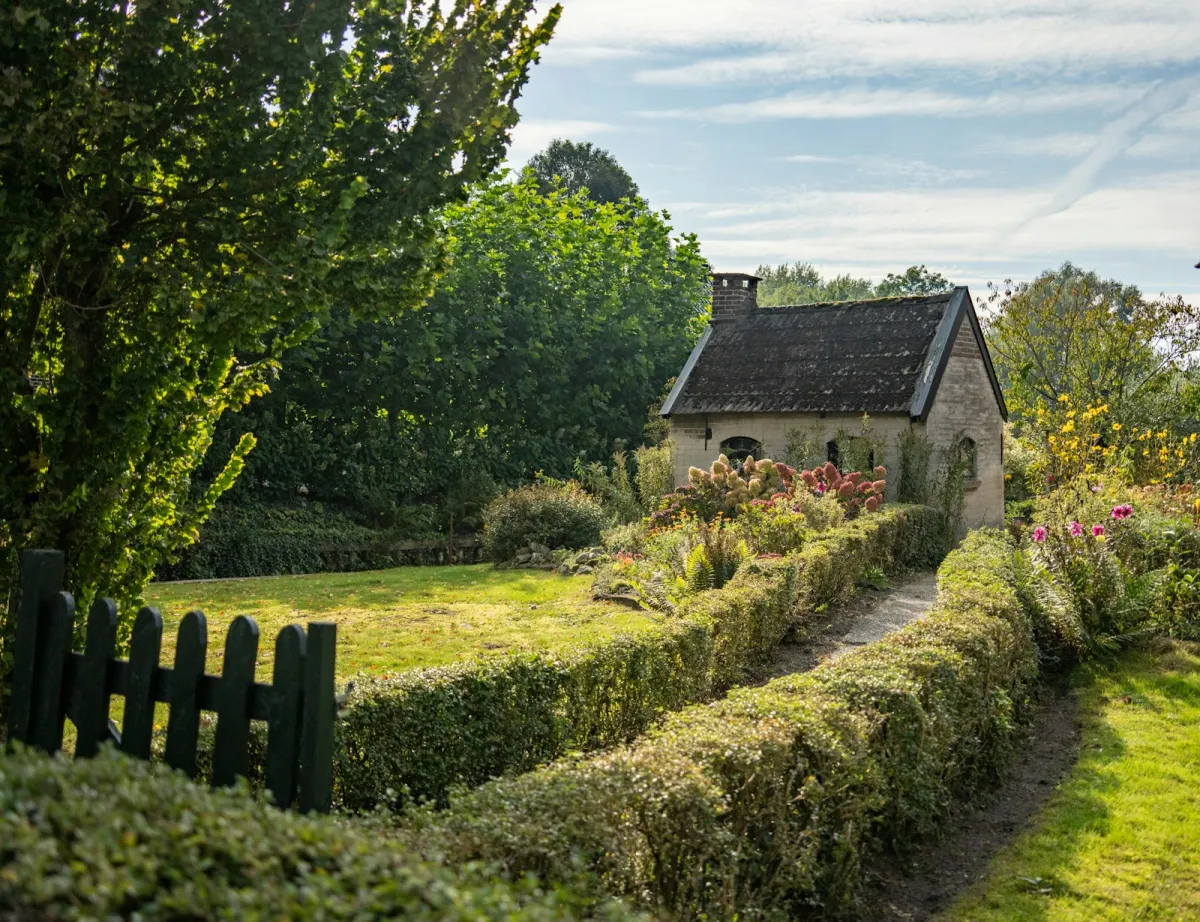 A small brick cottage behind a hedge-lined path and a flower garden