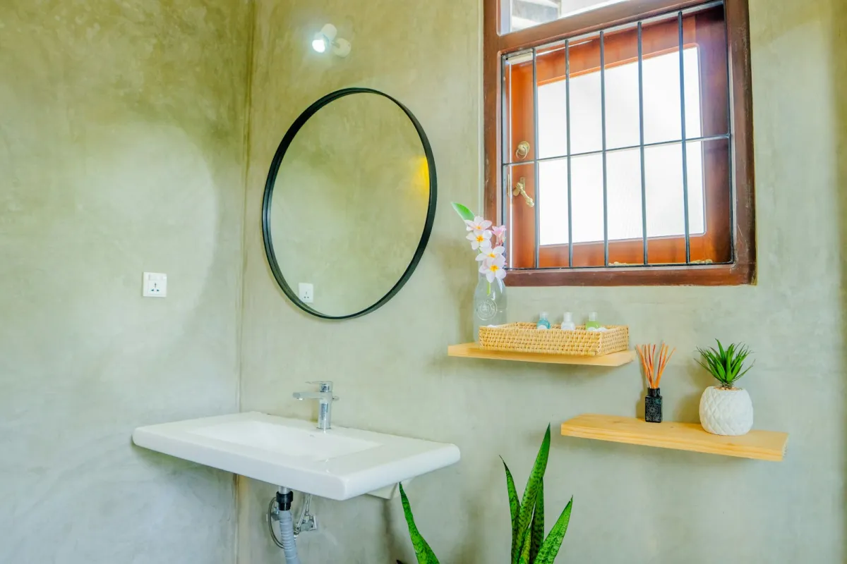 Bright bathroom with a round mirror, white basin and wooden shelves holding plants and toiletries