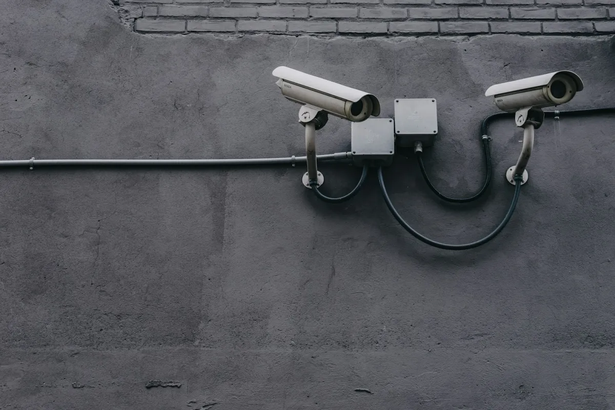 Two white security cameras mounted on a grey wall, linked by black cables and junction boxes Two white security cameras mounted on a grey wall, linked by black cables and junction boxes