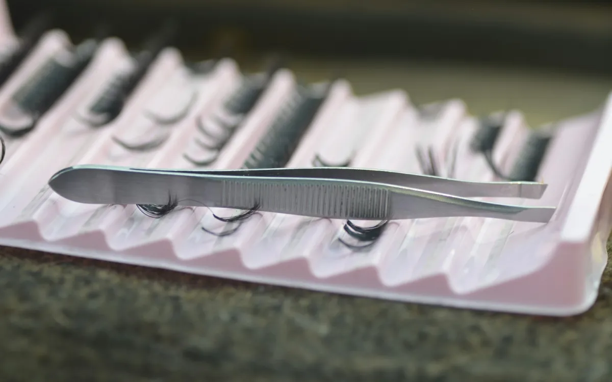 Stainless steel tweezers resting on a pink tray of false eyelash clusters Stainless steel tweezers resting on a pink tray of false eyelash clusters