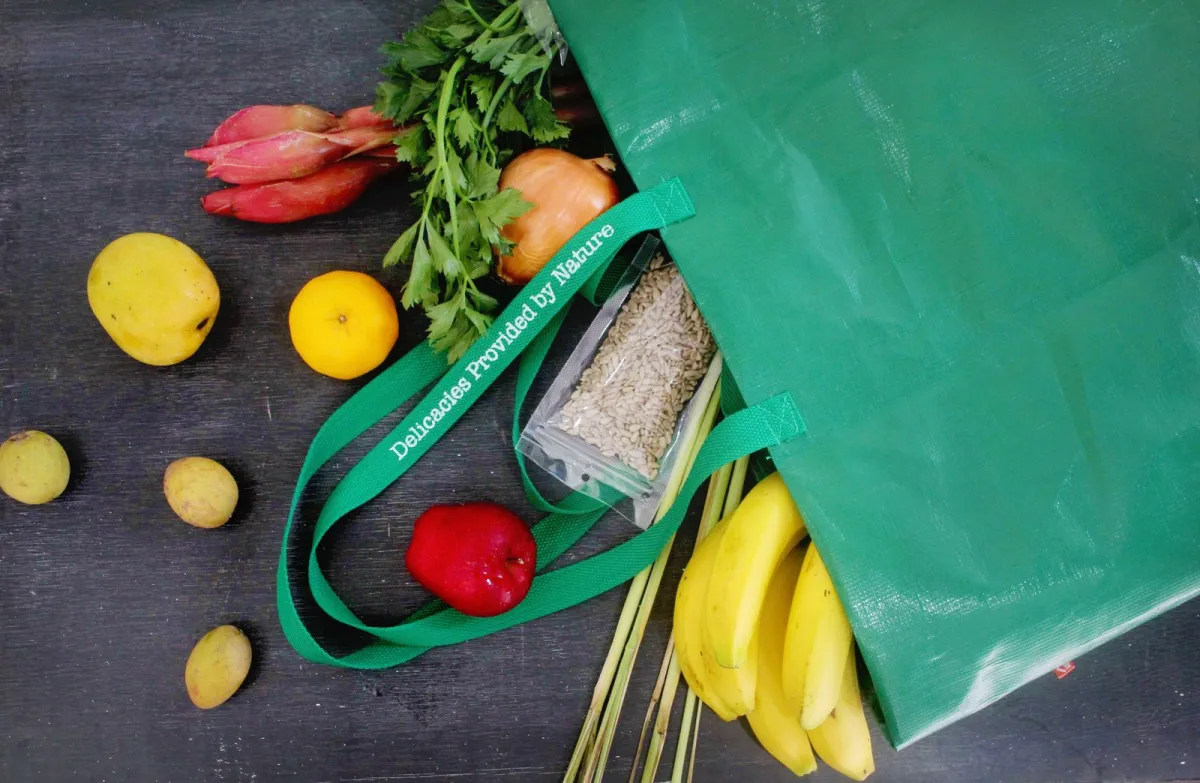 Fresh fruit, vegetables, herbs and bananas spilling out of a green reusable shopping bag onto a dark table Fresh fruit, vegetables, herbs and bananas spilling out of a green reusable shopping bag onto a dark table