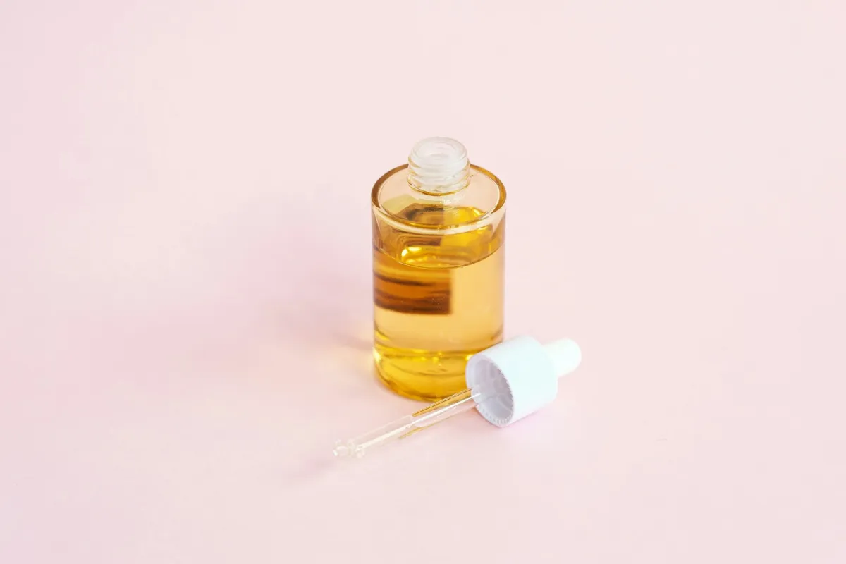 An open bottle of golden face oil with its dropper on a pale pink surface An open bottle of golden face oil with its dropper on a pale pink surface