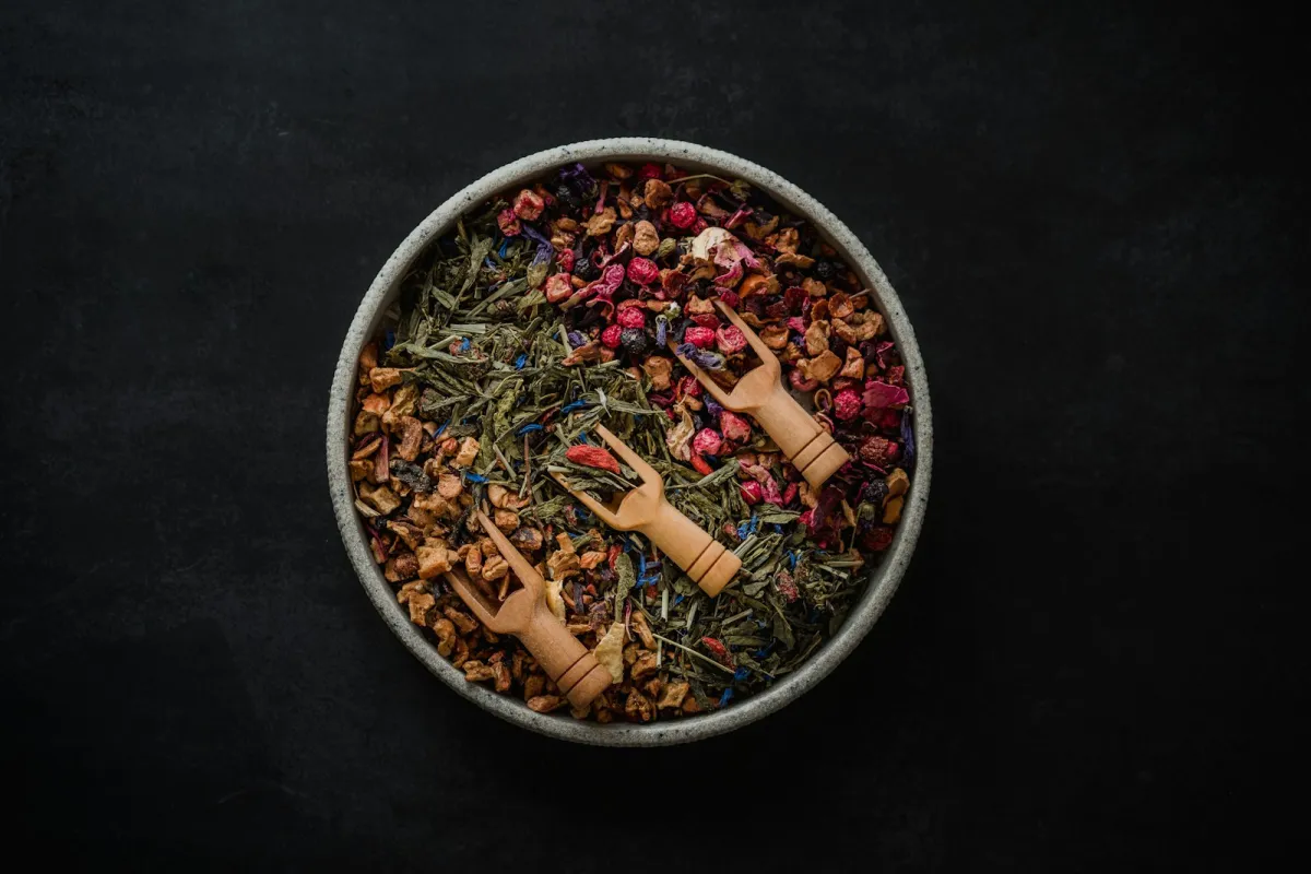 Bowl of loose herbal tea blends with three small wooden scoops on a dark background