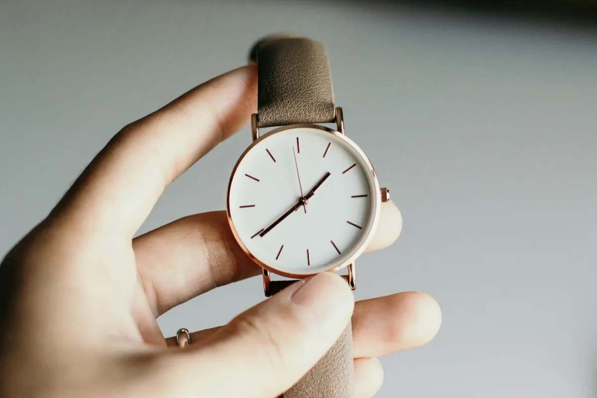 A hand holding up a minimalist white-faced watch with a rose gold case and taupe leather strap