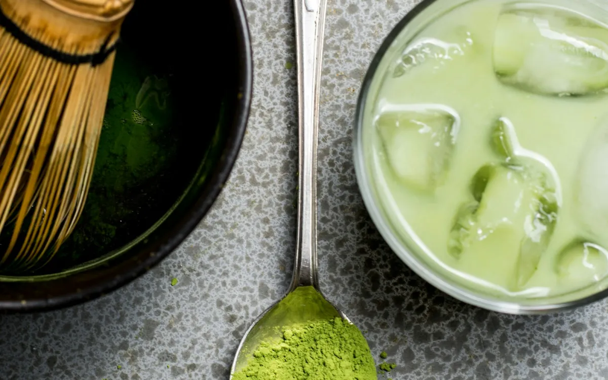 A spoonful of green powder beside an iced green drink and a bamboo whisk A spoonful of green powder beside an iced green drink and a bamboo whisk