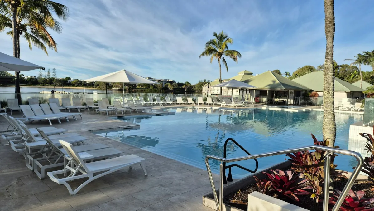 A resort swimming pool surrounded by white sun loungers, parasols and palm trees under a blue sky