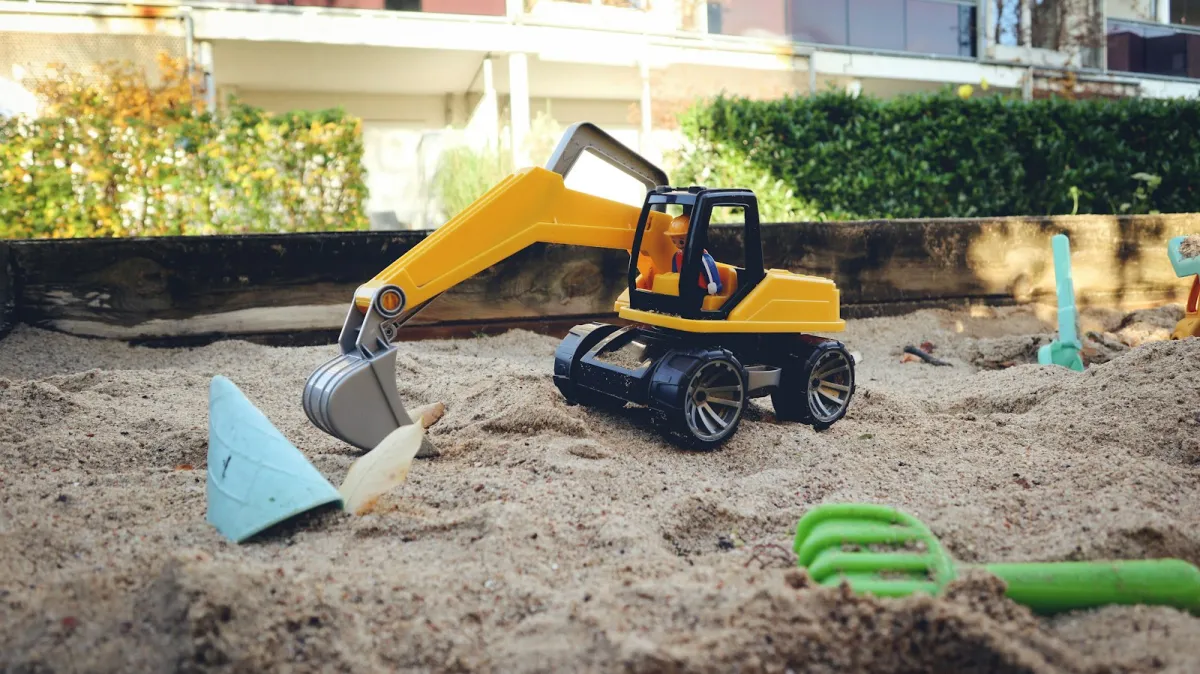 A yellow toy digger in a garden sandpit with spades and a rake A yellow toy digger in a garden sandpit with spades and a rake