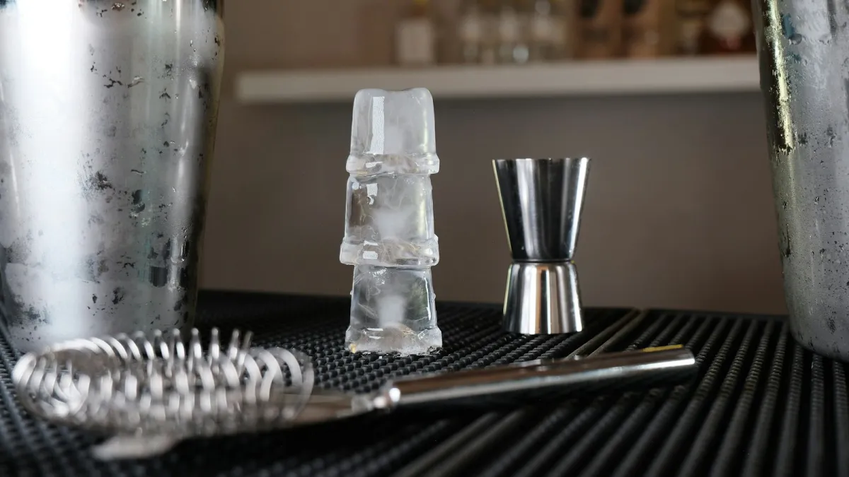 A cocktail shaker, jigger, strainer and stacked ice cubes on a bar mat