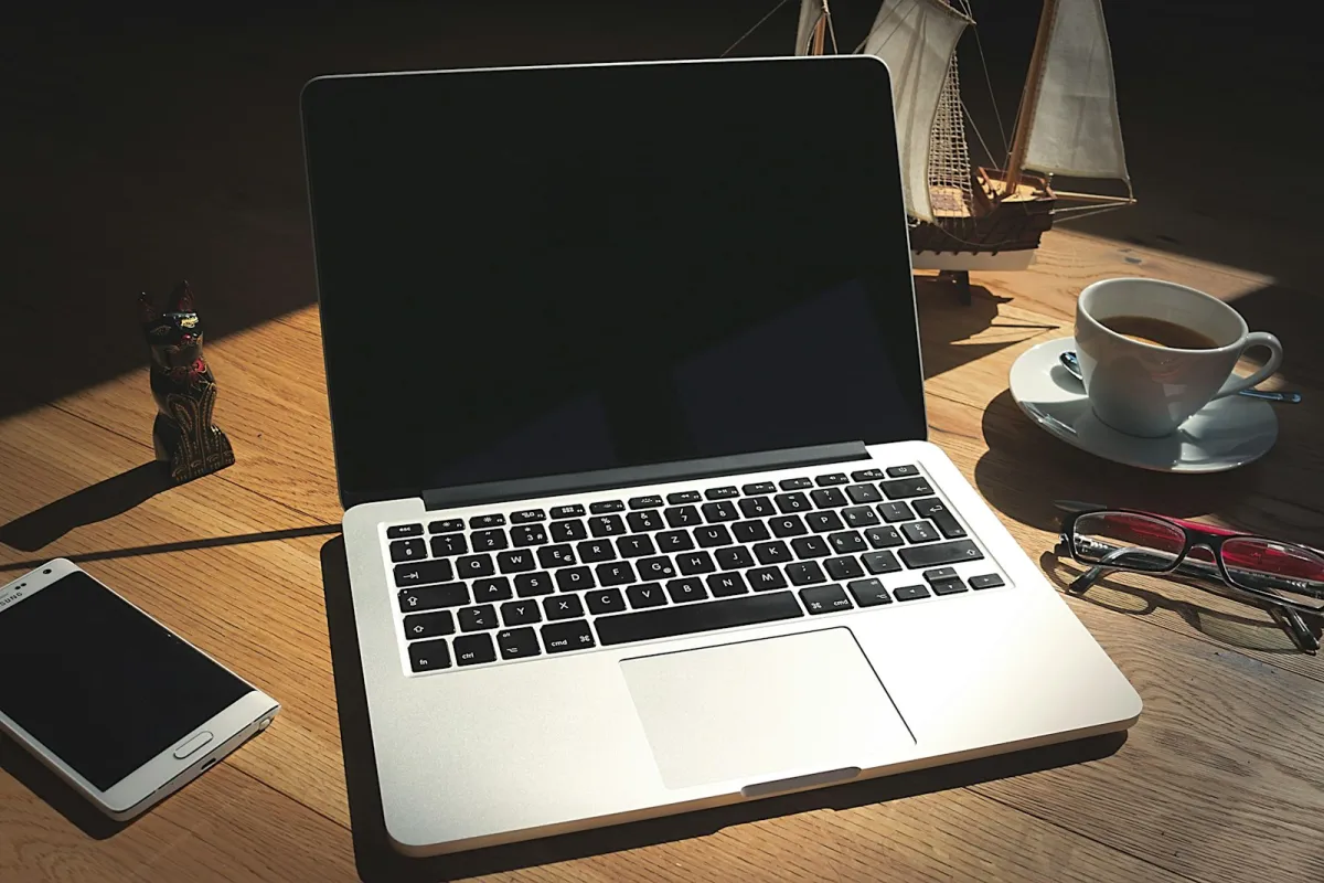 An open laptop on a wooden desk beside a smartphone, a cup of coffee and a pair of glasses An open laptop on a wooden desk beside a smartphone, a cup of coffee and a pair of glasses