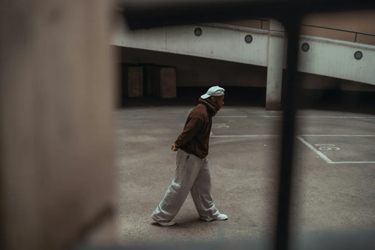 Person in a brown hoodie, cap and grey joggers walking across an empty concrete space