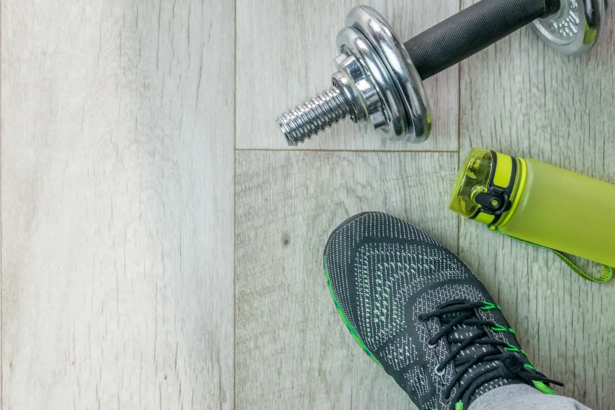 A trainer, a dumbbell and a green water bottle on a light wooden floor