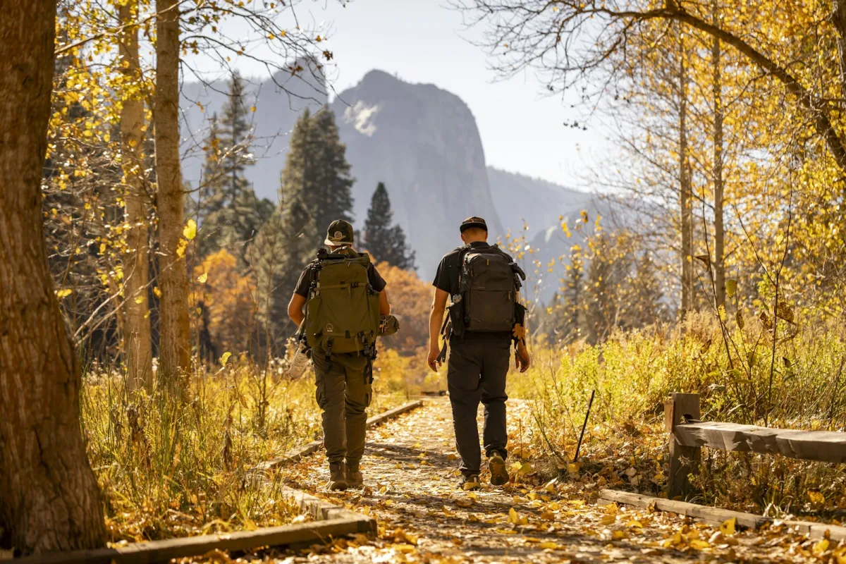 Two hikers with backpacks walking a leaf-covered trail through autumn woods towards a granite cliff