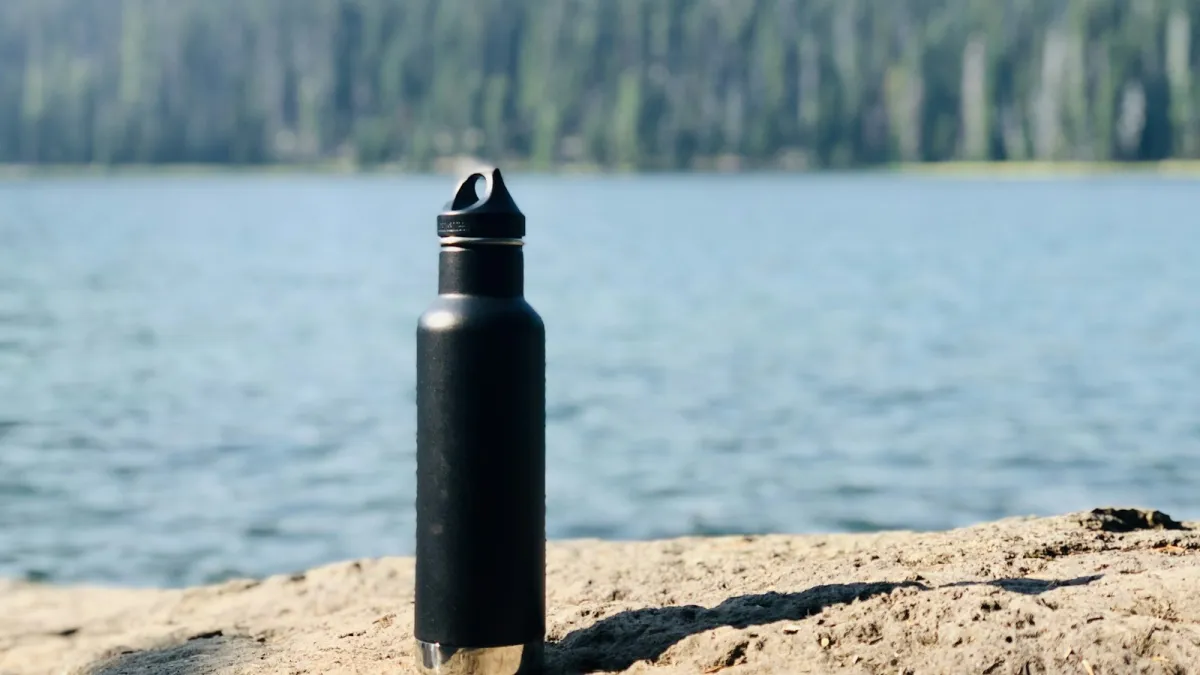 A black reusable water bottle standing on a rock beside a lake with pine forest behind