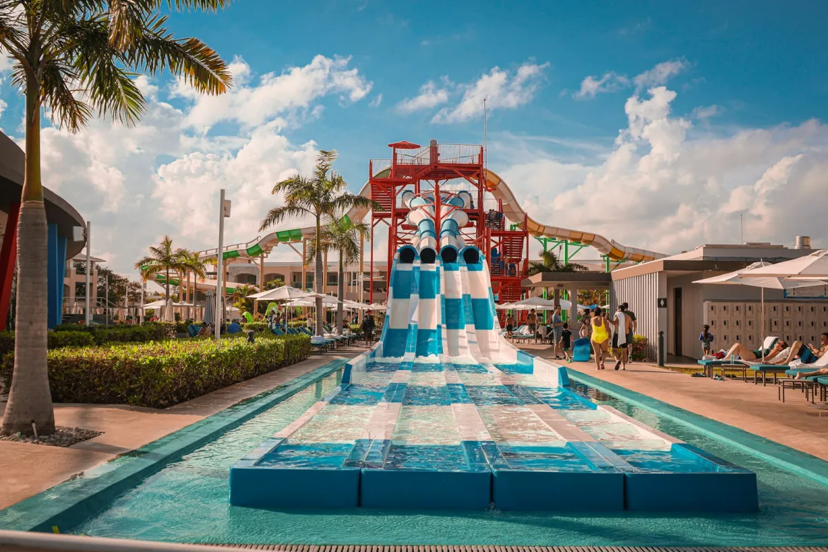 A blue and white multi-lane water slide splashing into a pool, with palm trees and a slide tower behind