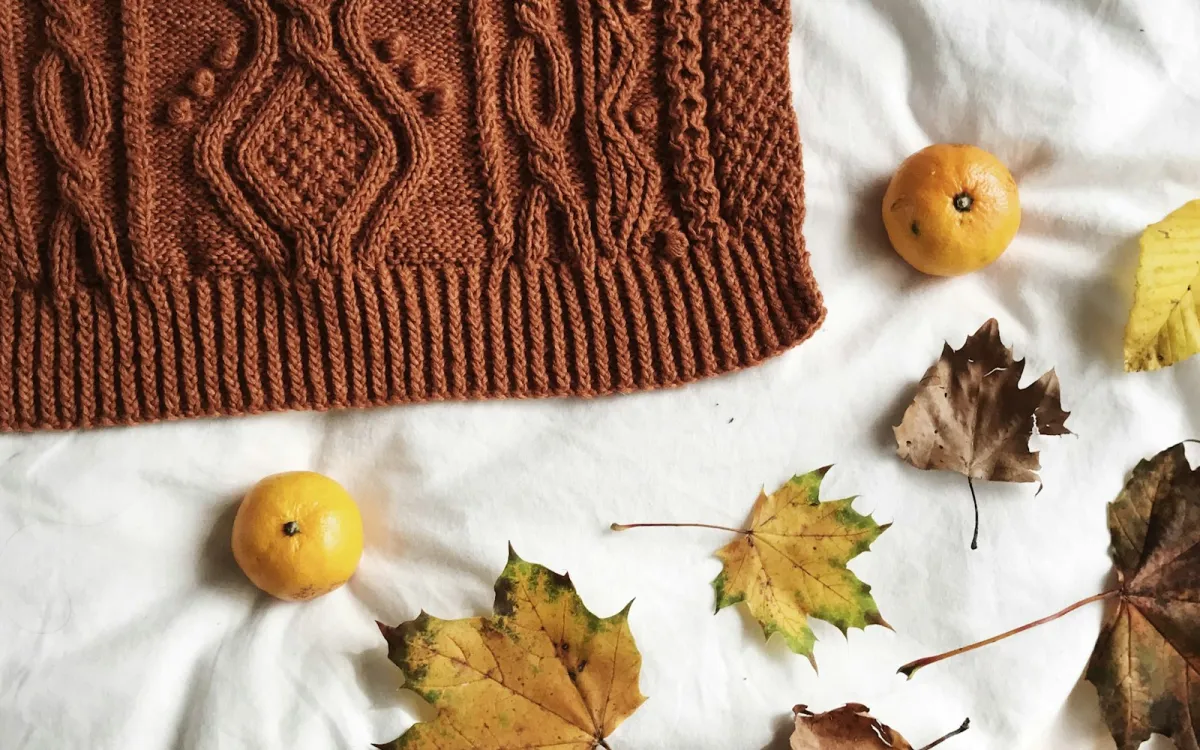 A rust-brown cable-knit jumper on a white sheet with autumn leaves and two clementines