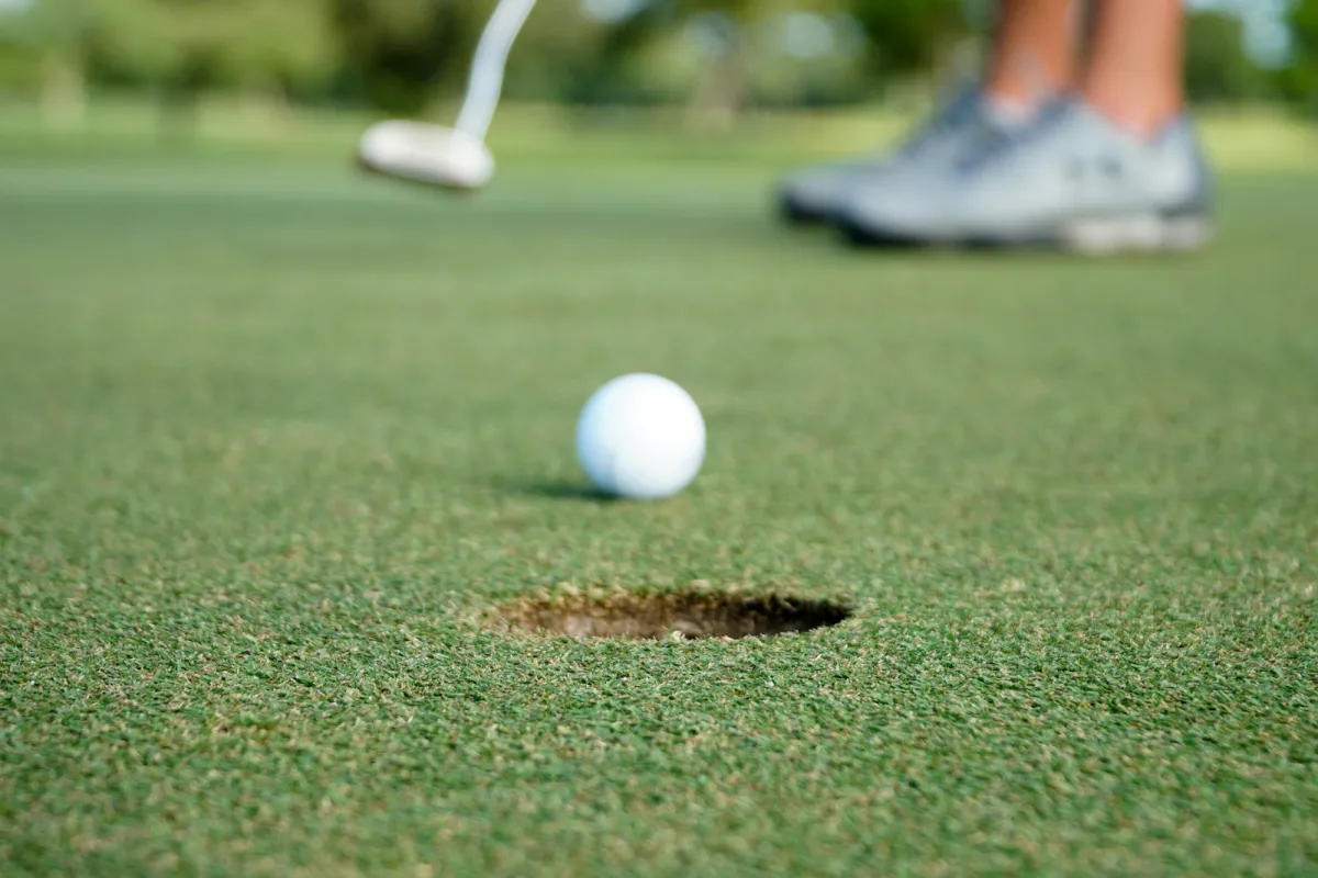 Golf ball rolling towards the hole on a putting green, with a putter and golf shoes blurred behind