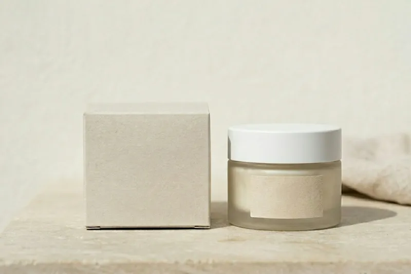 A frosted glass cream jar with a white lid beside a plain cardboard box