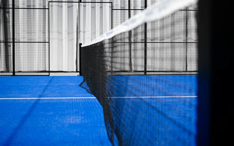 Low view along the net of an empty indoor padel court with a blue playing surface