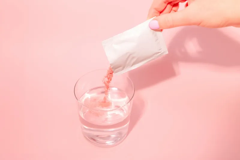 A hand pouring pink powder from a sachet into a glass of water on a pink background